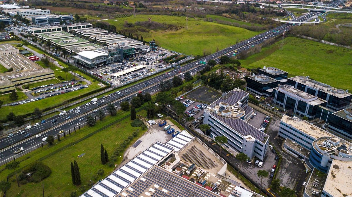 Aerial view of a wide Italian highway with three lanes in each direction. So many cars, motorcycles, trucks are running on this road near a big city and an industrial area.